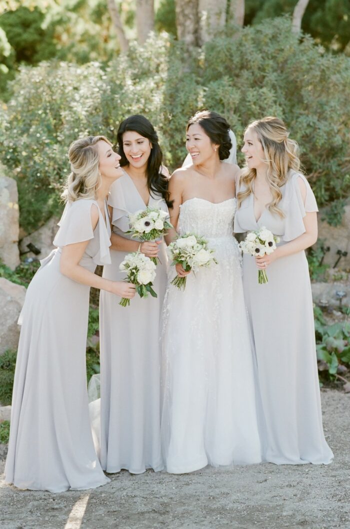 Bride with bridesmaids laughing with each other holding bouquet and wearing white dresses