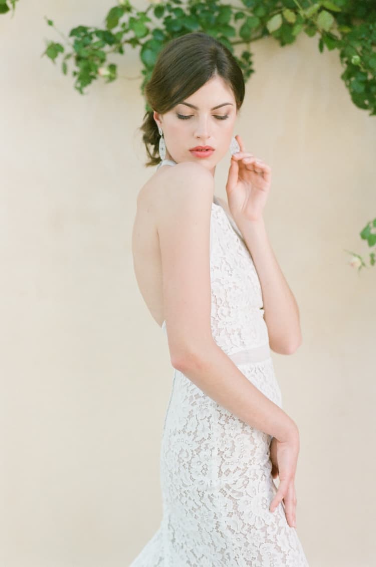 Bride in her lace dress standing in front of a yellow stonewall touching her earring