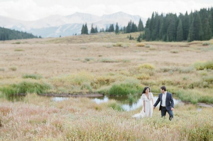 Far out shot of engaged couple in tall grass next to water and mountain backdrop