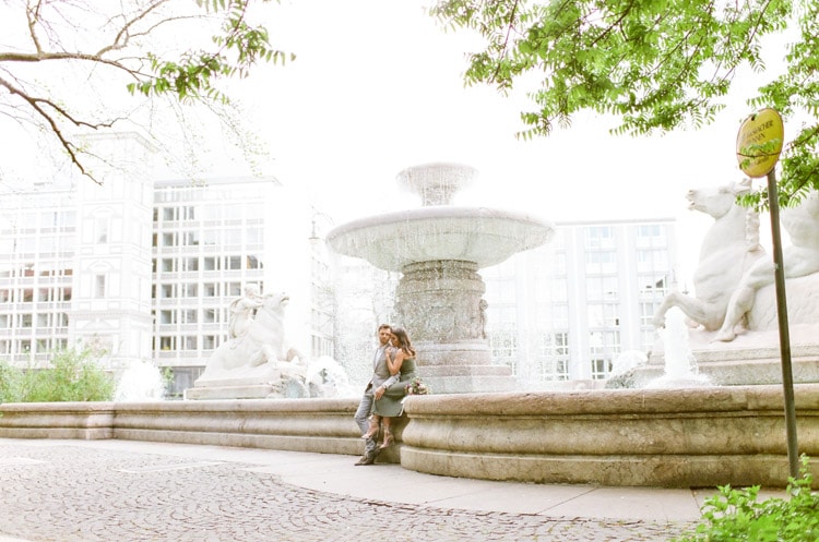 Couple sitting in front of fountain in Munich during their destination engagement portrait session