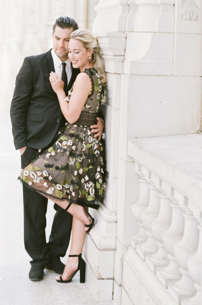 Couple leaning against white stone wall wearing black suit and black floral mini dress