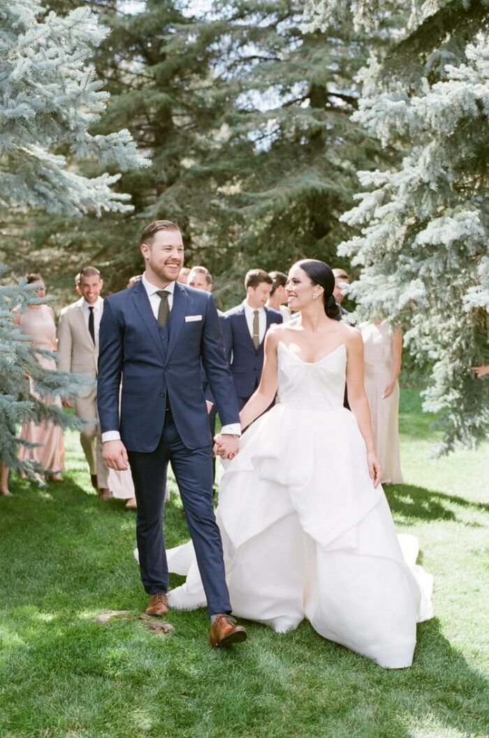 Groom wearing navy blue custom suit holding hands with bride in white wedding gown and backdrop of pine trees