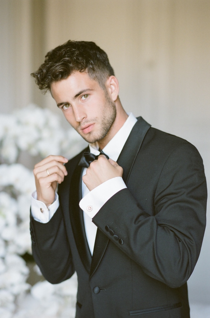 Stylish groom in tuxedo laughs suit marsala red, burgundy bow tie. Man  buttoning his jacket outdoors. Professional hairstyle, beard, mustache.  Wedding preparations, getting ready. Copy space for text. - Stock Image -  Everypixel